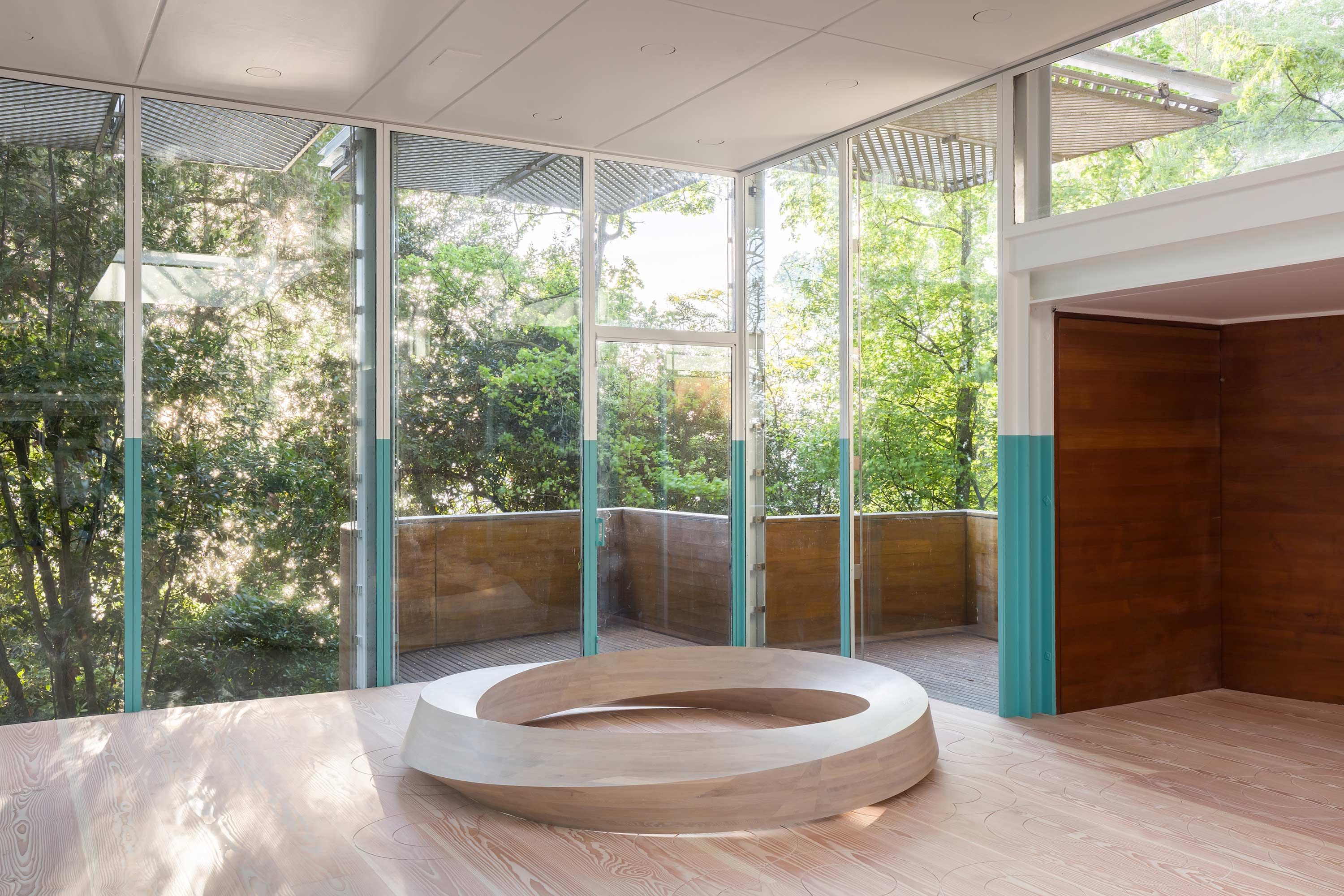 A spacious room with floor-to-ceiling glass walls revealing a green landscape, with a large wooden ring sculpture on the f...