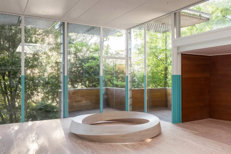 A spacious room with floor-to-ceiling glass walls revealing a green landscape, with a large wooden ring sculpture on the f...
