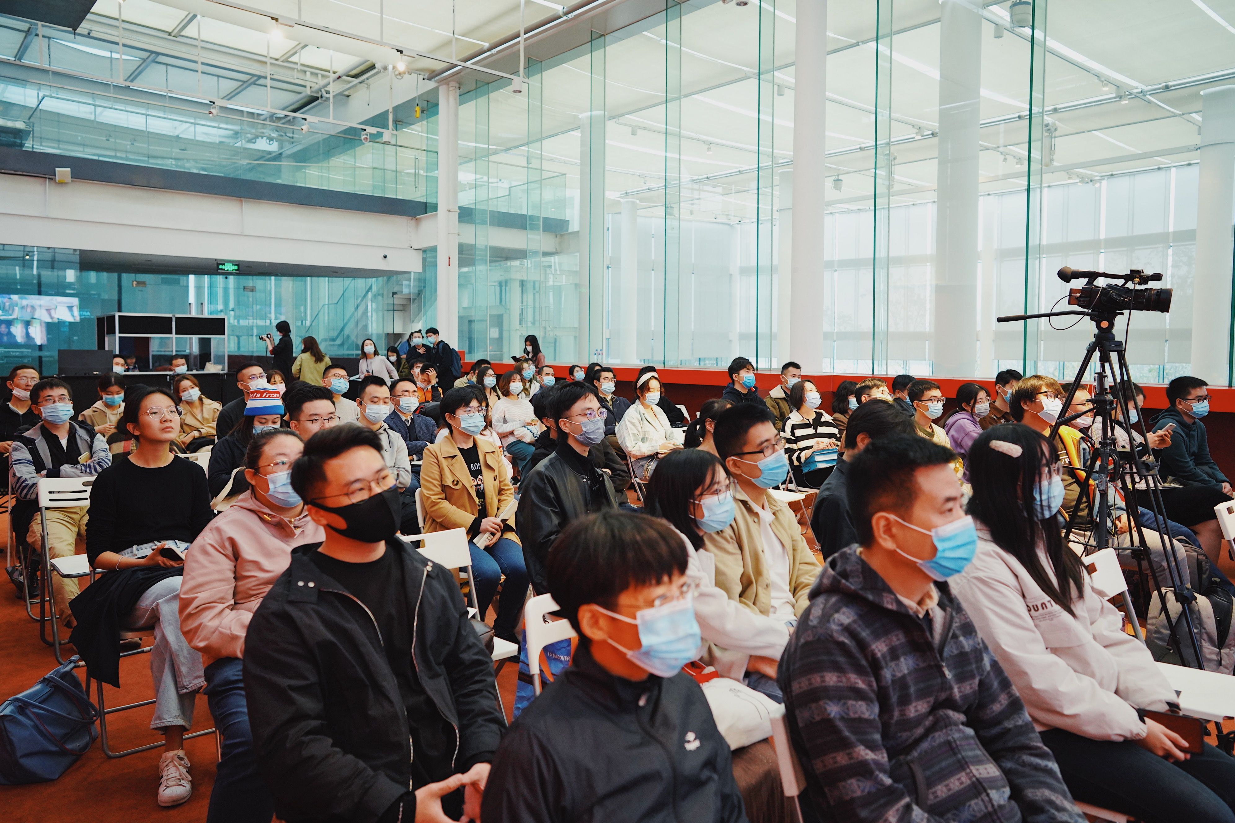 An audience seated on various chairs in a well-organized view, attentively listening to off-screen proceedings. A lone cam...