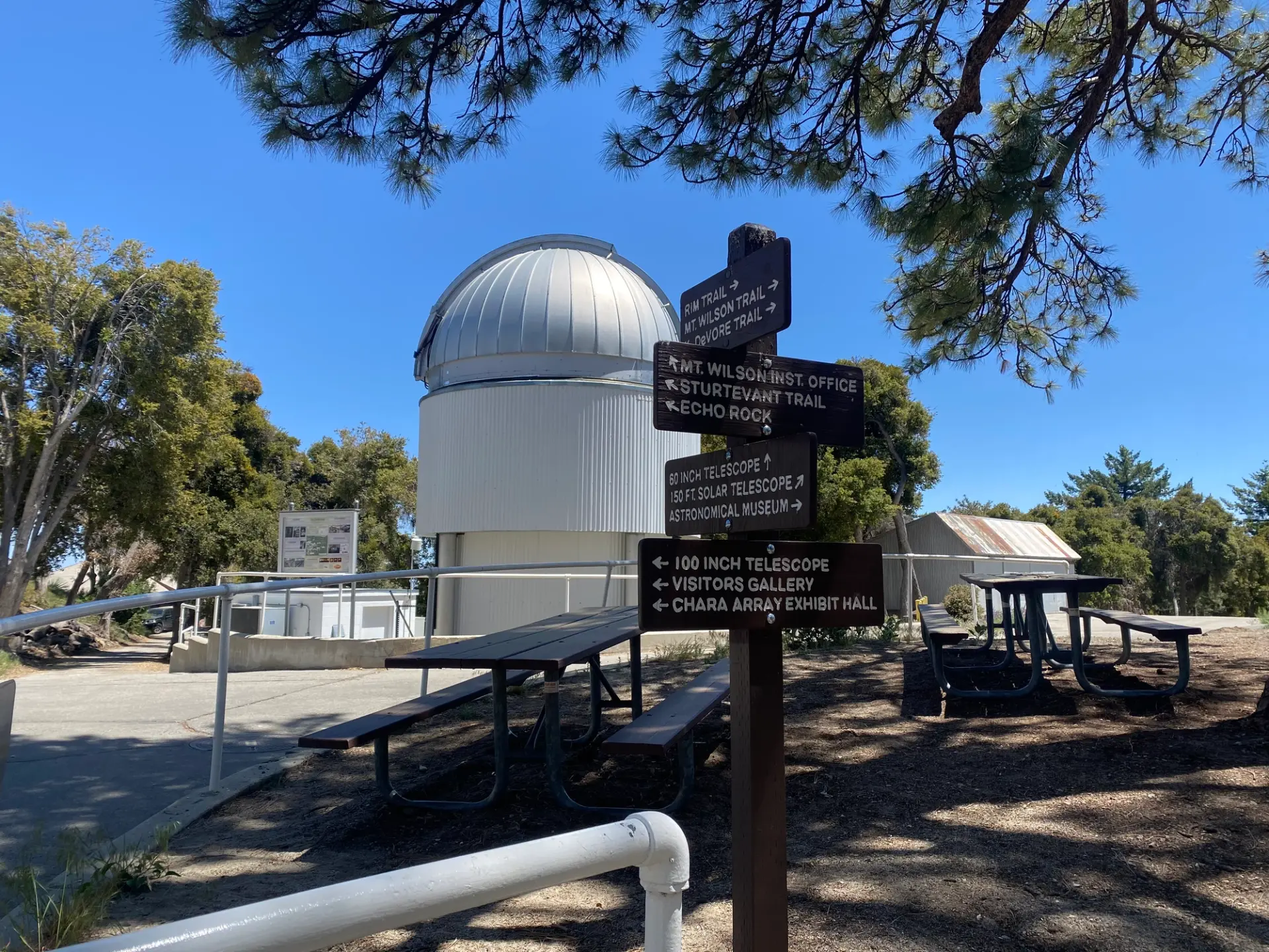 A wooden direction signpost in the foreground stands against the backdrop of a large white cylindrical building containing a space telescope.