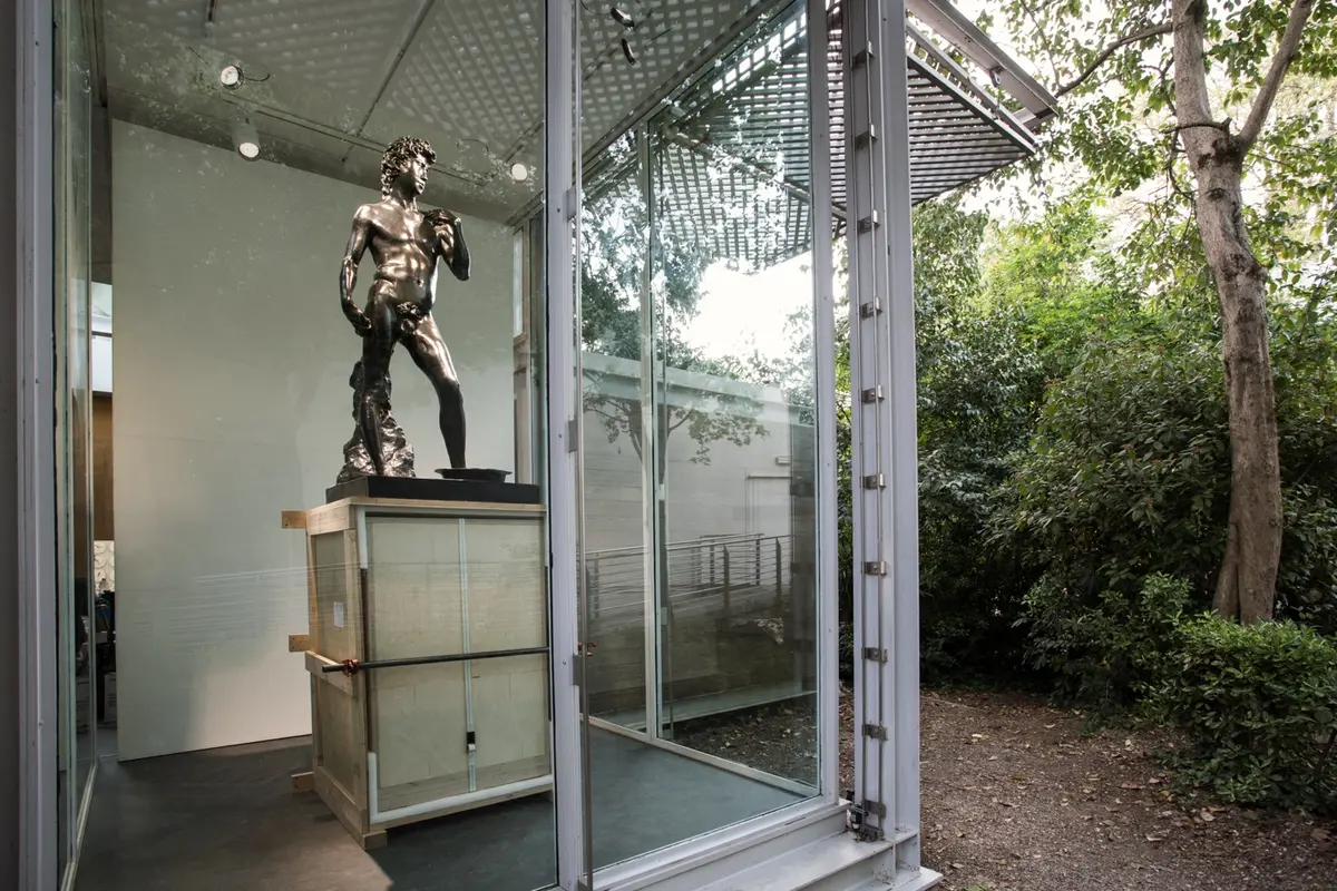 A black glossy statue of a naked figure displayed in a glass-walled gallery, with the lush greenery of the outside environment visible in the background.