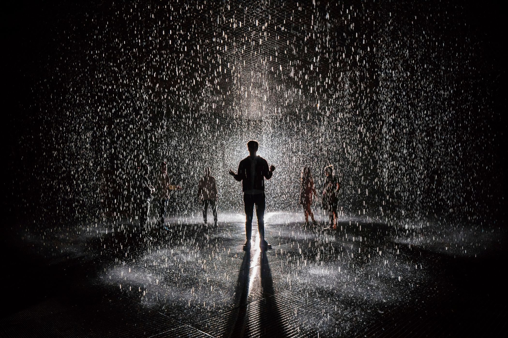 A silhouette of a person in the spotlight amidst heavy rain against a dark background, surrounded by multiple other vague ...