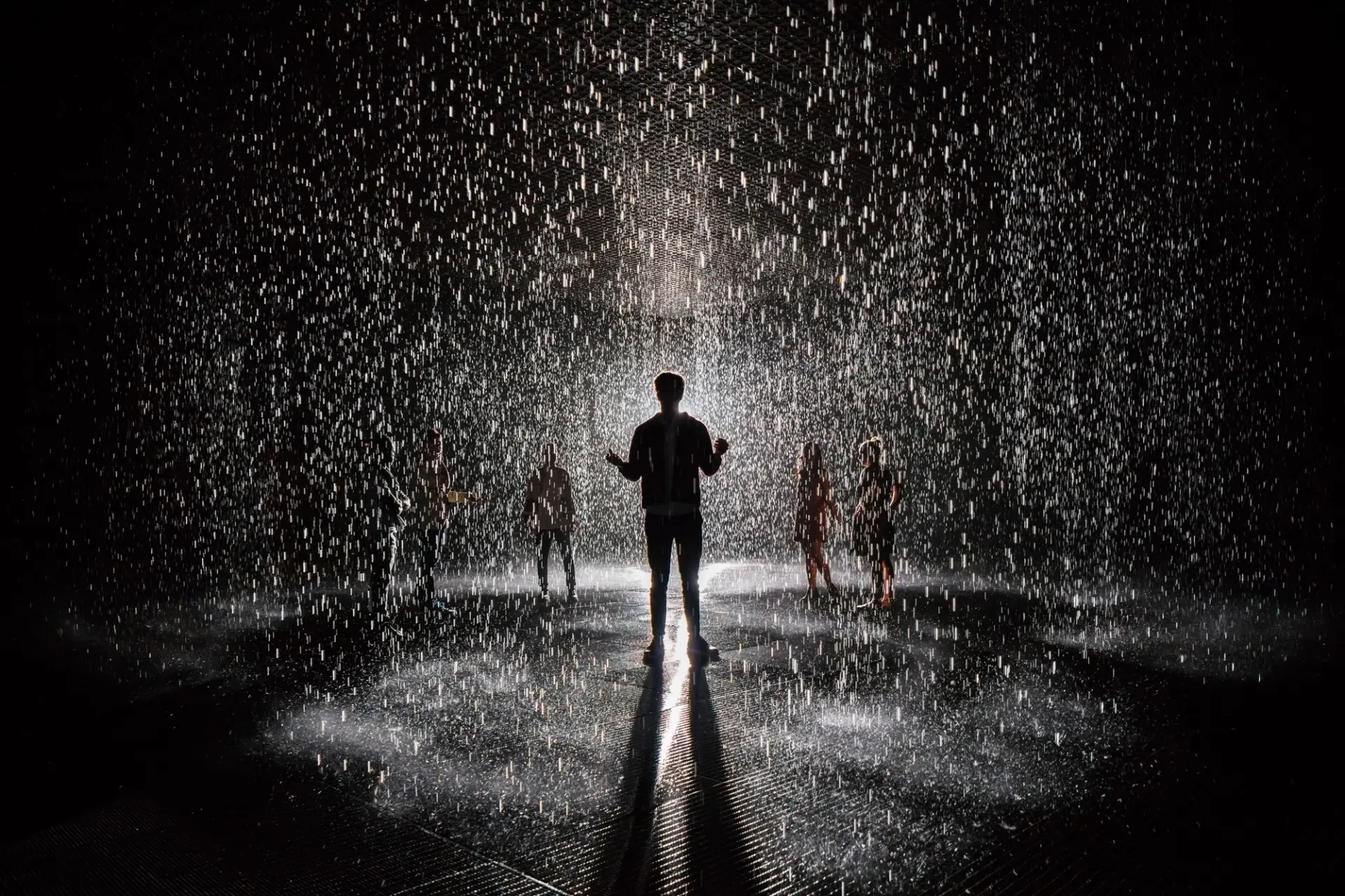 A silhouette of a person in the spotlight amidst heavy rain against a dark background, surrounded by multiple other vague ...