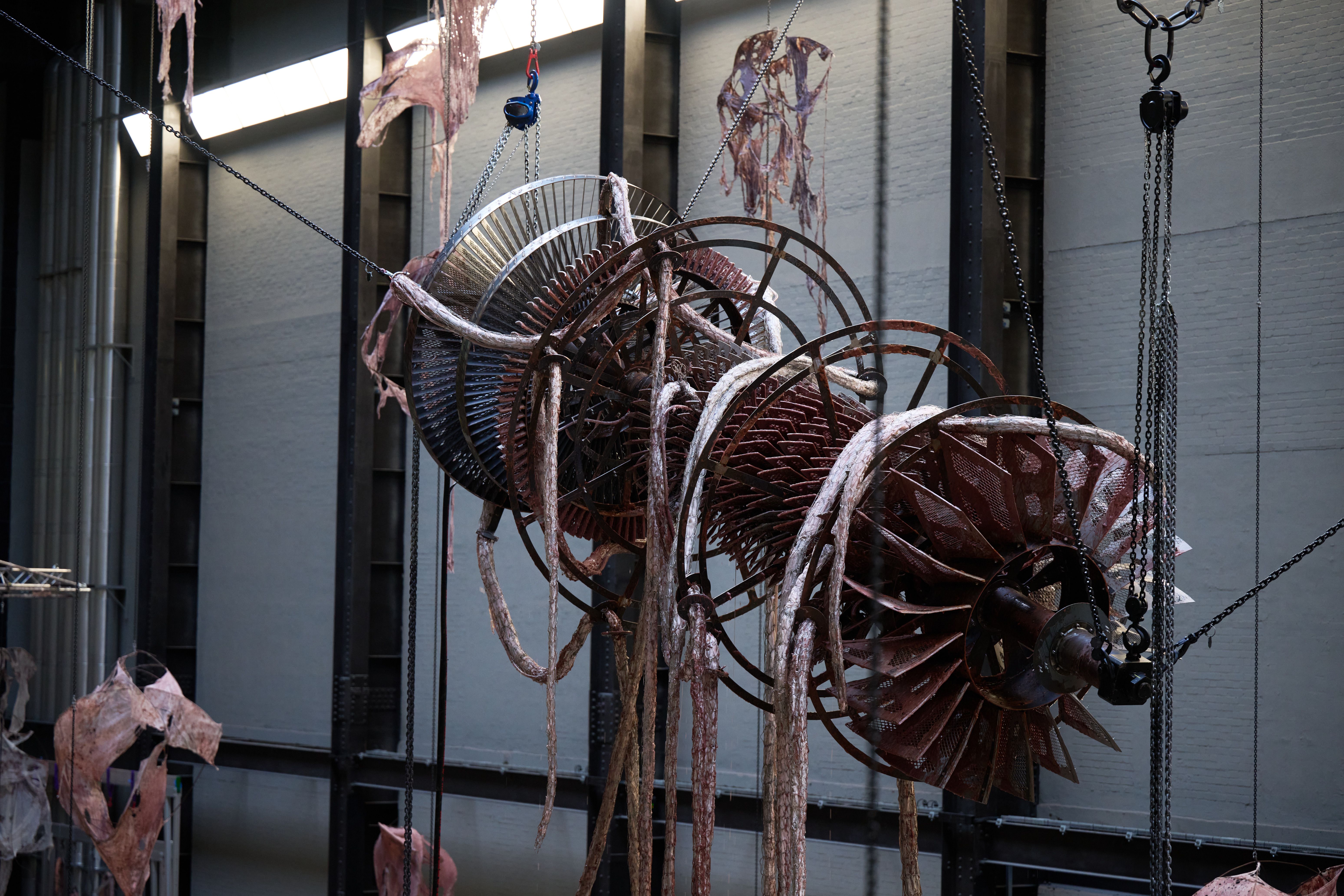 Detailed view of a large mechanical structure entwined with fleshy material, suspended by chains.