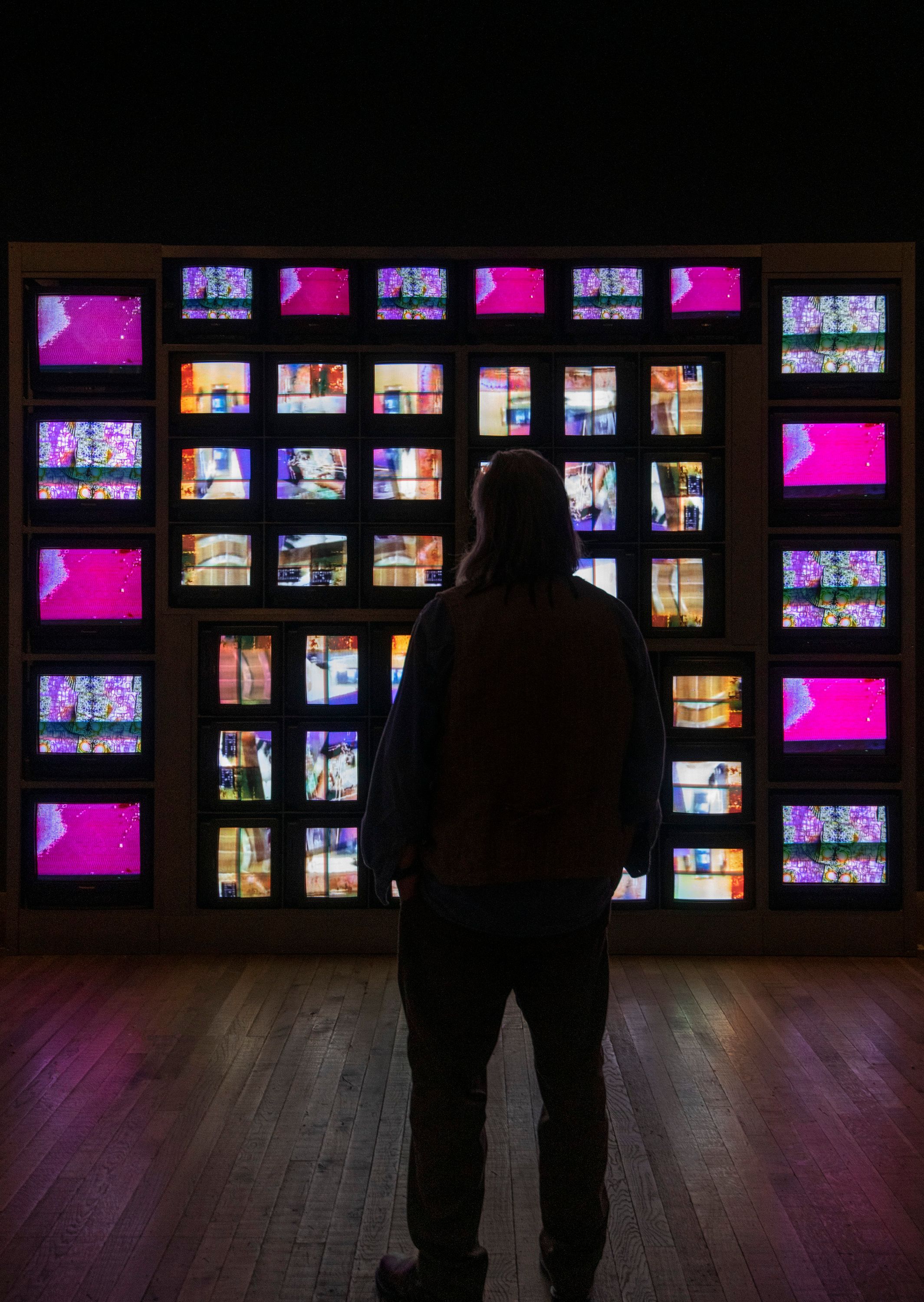 A silhouette of a person stands observing a large wall filled with numerous monitors, each displaying vibrant abstract ima...
