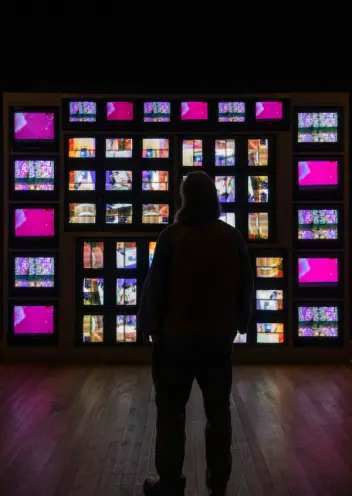 A silhouette of a person stands observing a large wall filled with numerous monitors, each displaying vibrant abstract ima...