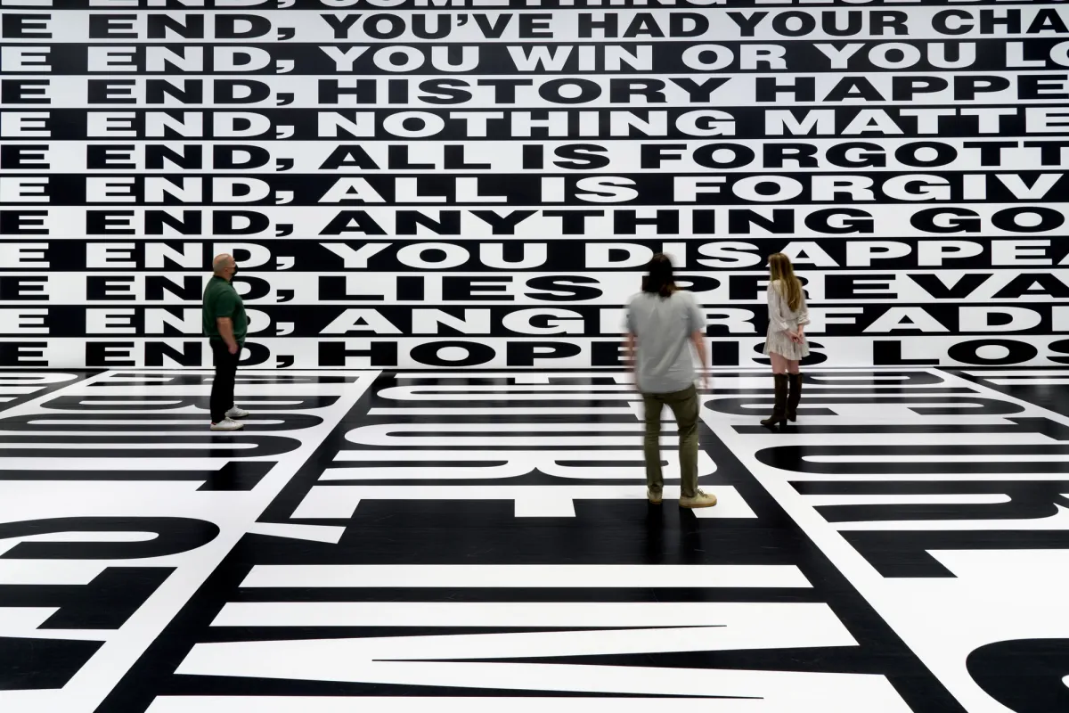 Three individuals stand in a vast room, completely enveloped by large-scale, black and white text that coats the walls and flooring.