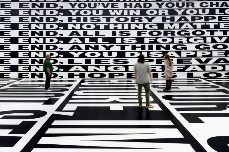 Three individuals stand in a vast room, completely enveloped by large-scale, black and white text that coats the walls and...