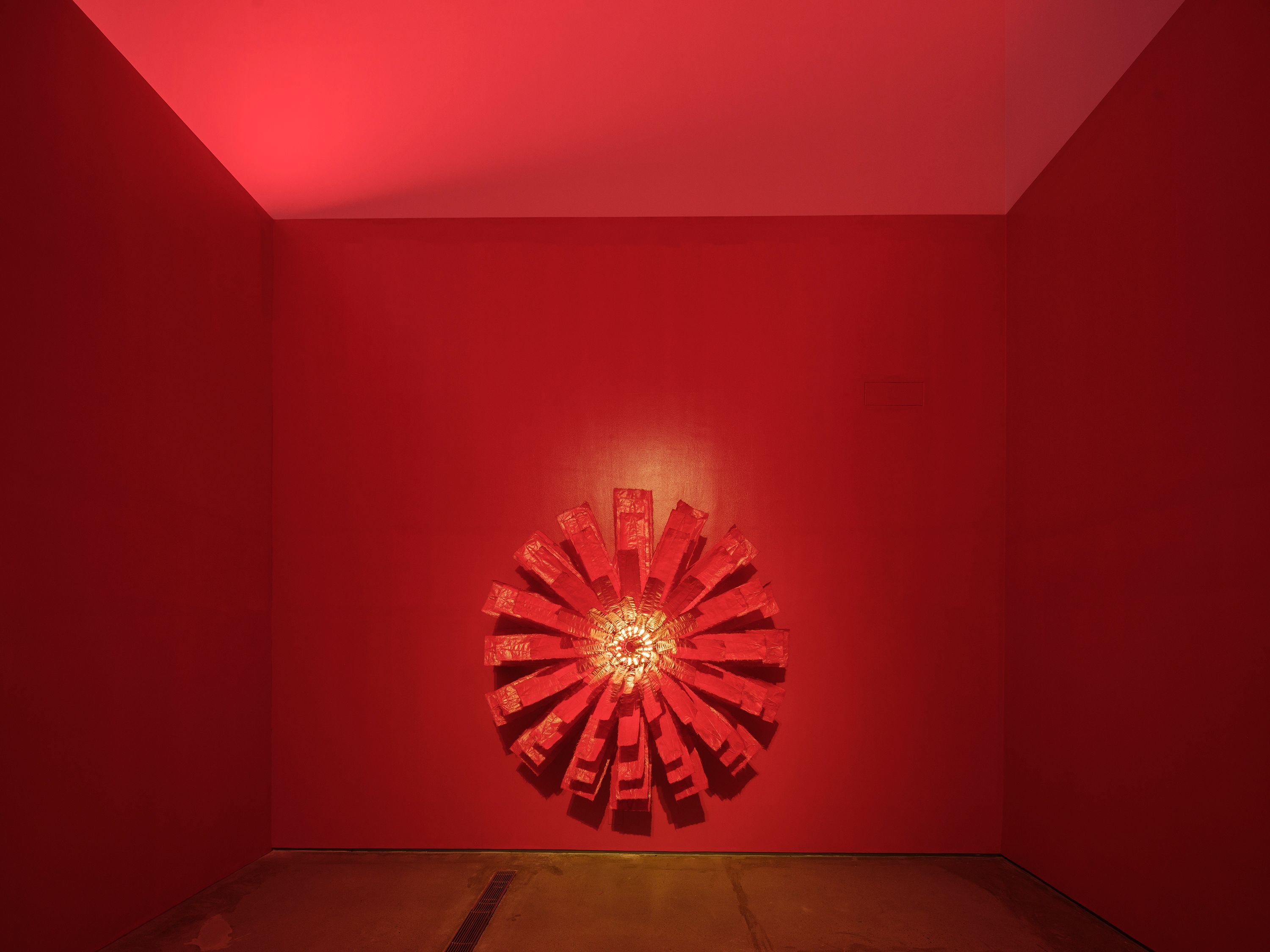 A vibrant, three-dimensional geometric artwork in a red tinted room. The sculpture, built from metal and acrylic on soft T...