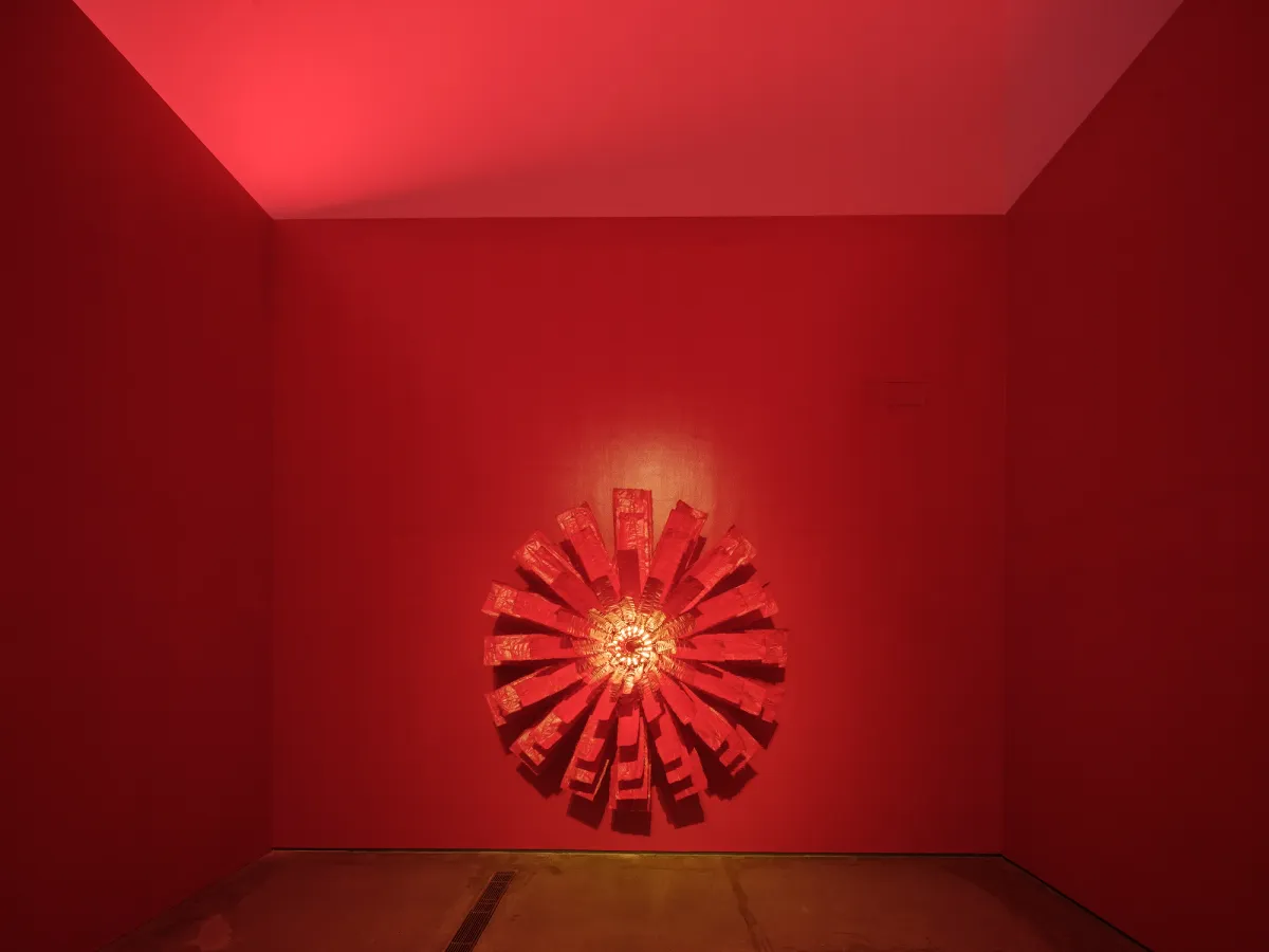 A vibrant, three-dimensional geometric artwork in a red tinted room. The sculpture, built from metal and acrylic on soft Tyvek, incorporates motorized components and LED elements.
