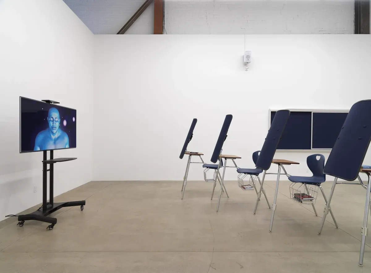 A screen positioned on a stand displays artwork in front of an gallery space decorated to look like a classroom. The room is lined with rows of blue chairs.