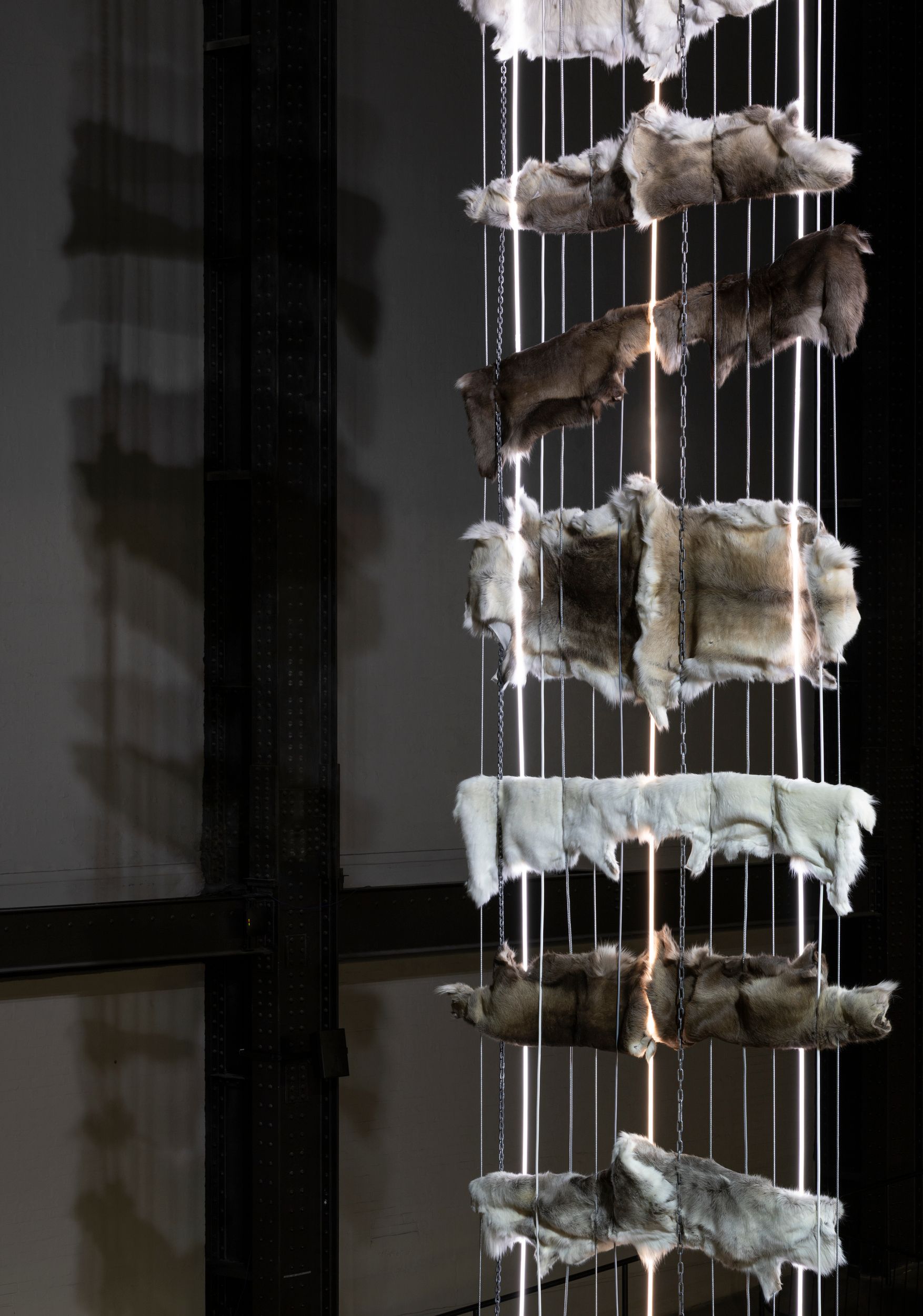 Vertically stacked animal hides hang in a tall column, threaded with chains and illuminated rods, inside a dark gallery space.