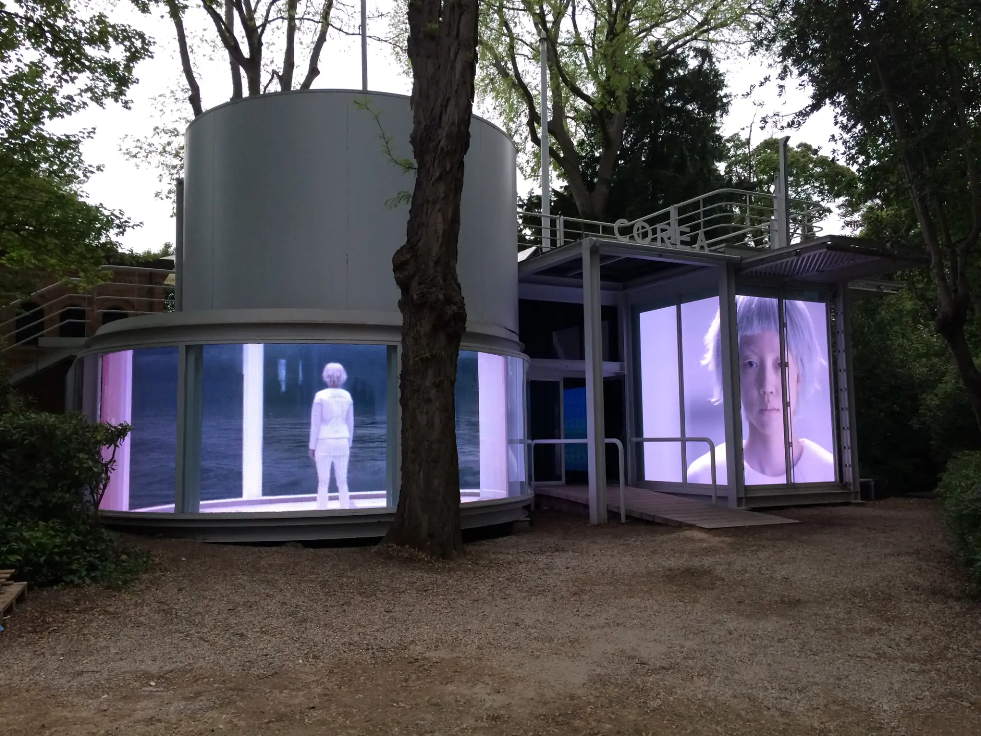 A hologram of a person dressed in white with white hair shown inside a cylindrical exhibition room with windowed walls. An...