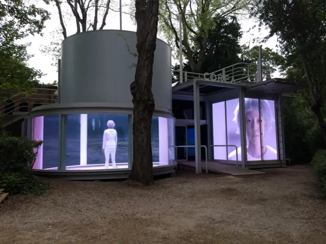 A hologram of a person dressed in white with white hair shown inside a cylindrical exhibition room with windowed walls. An...