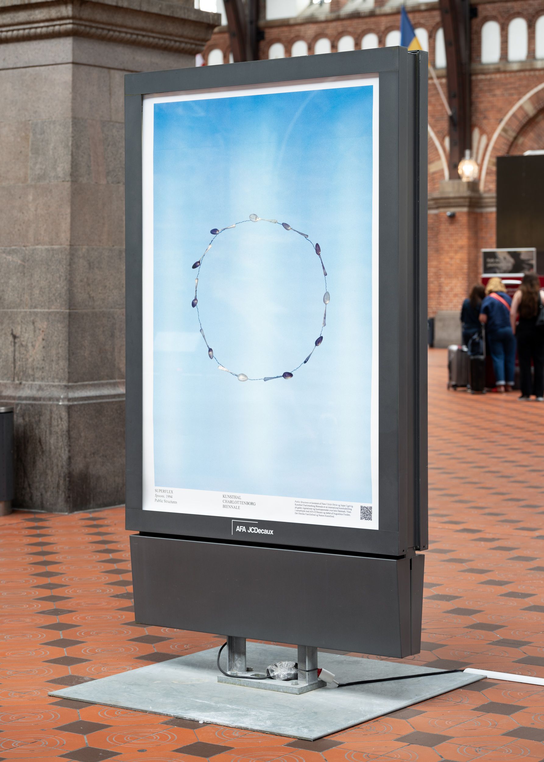 A small vertical billboard displaying an exhibition poster displaying a circle of eating spoons.