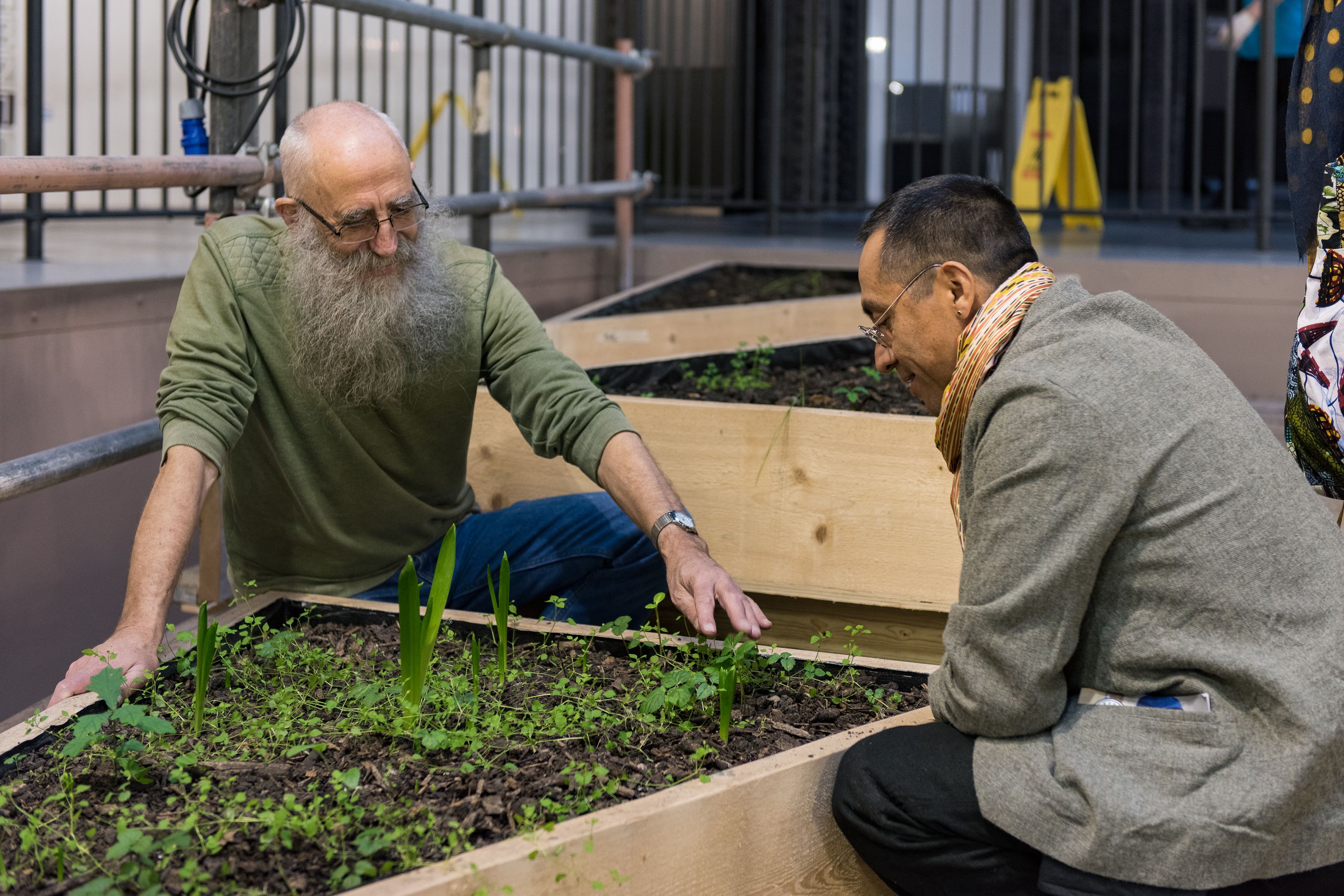 Two individuals sit next to a large, wooden, triangle-shaped planter box filled with sprouting leaves. Several other plant...
