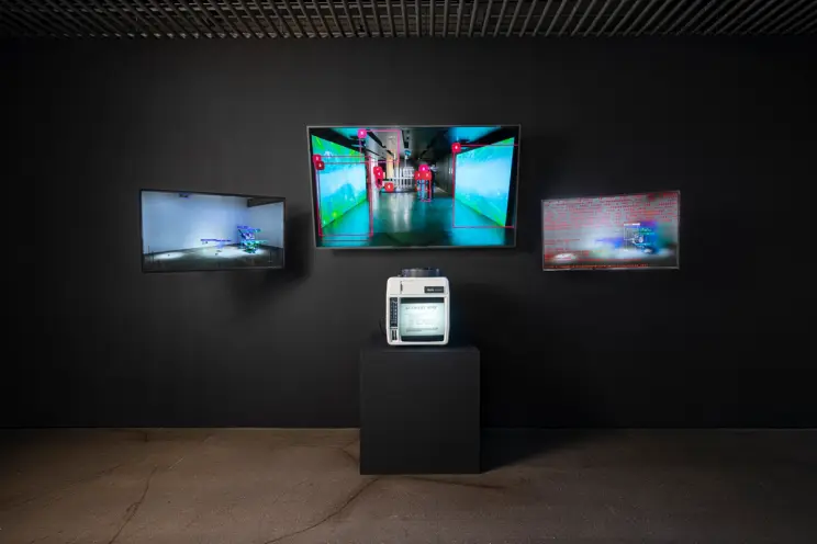 Three monitors displaying digital art are mounted on a black wall, with an electronic device placed on a black cubical str...