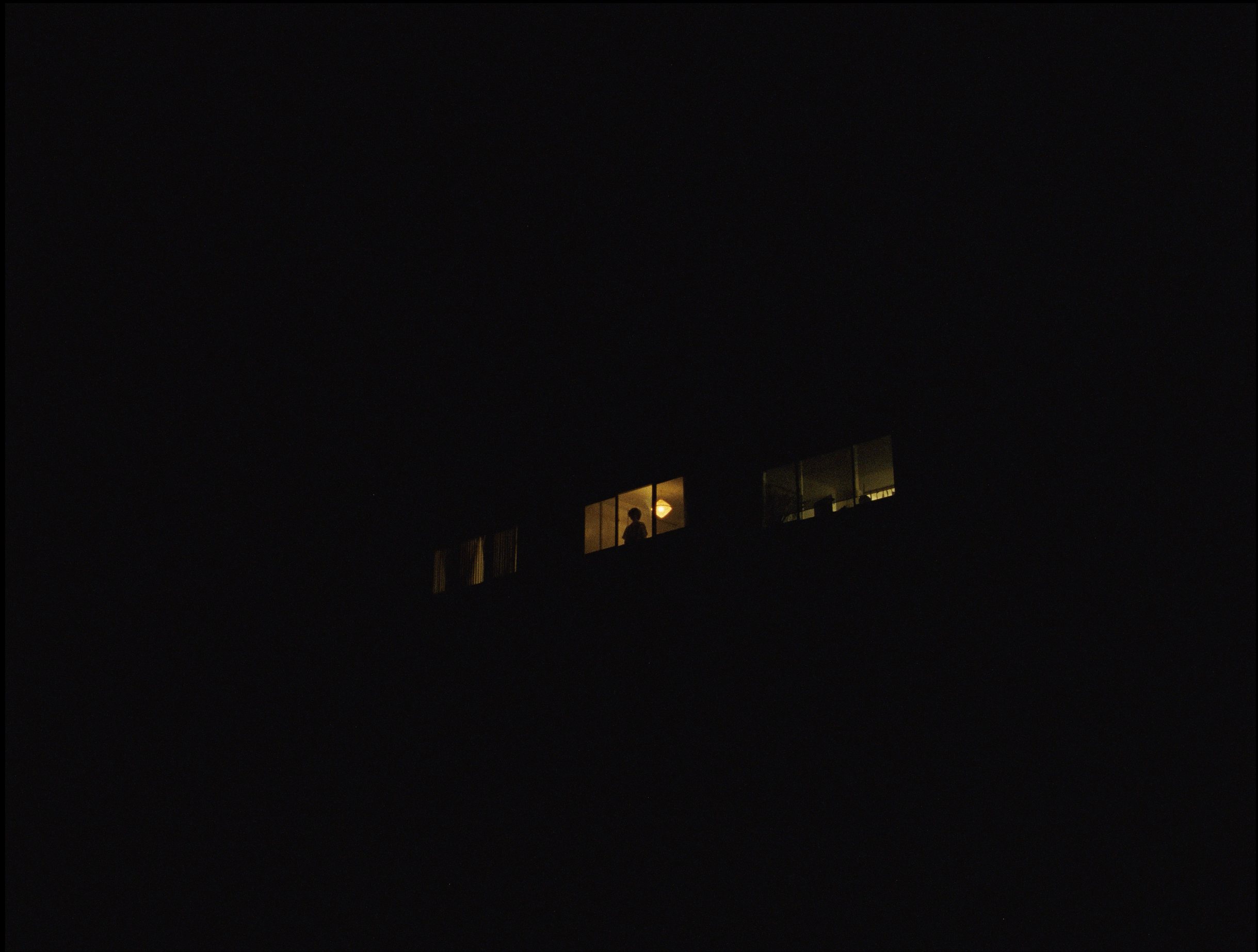 A distant window glowing with warm light in the darkness, showing a silhouette of a person standing inside.