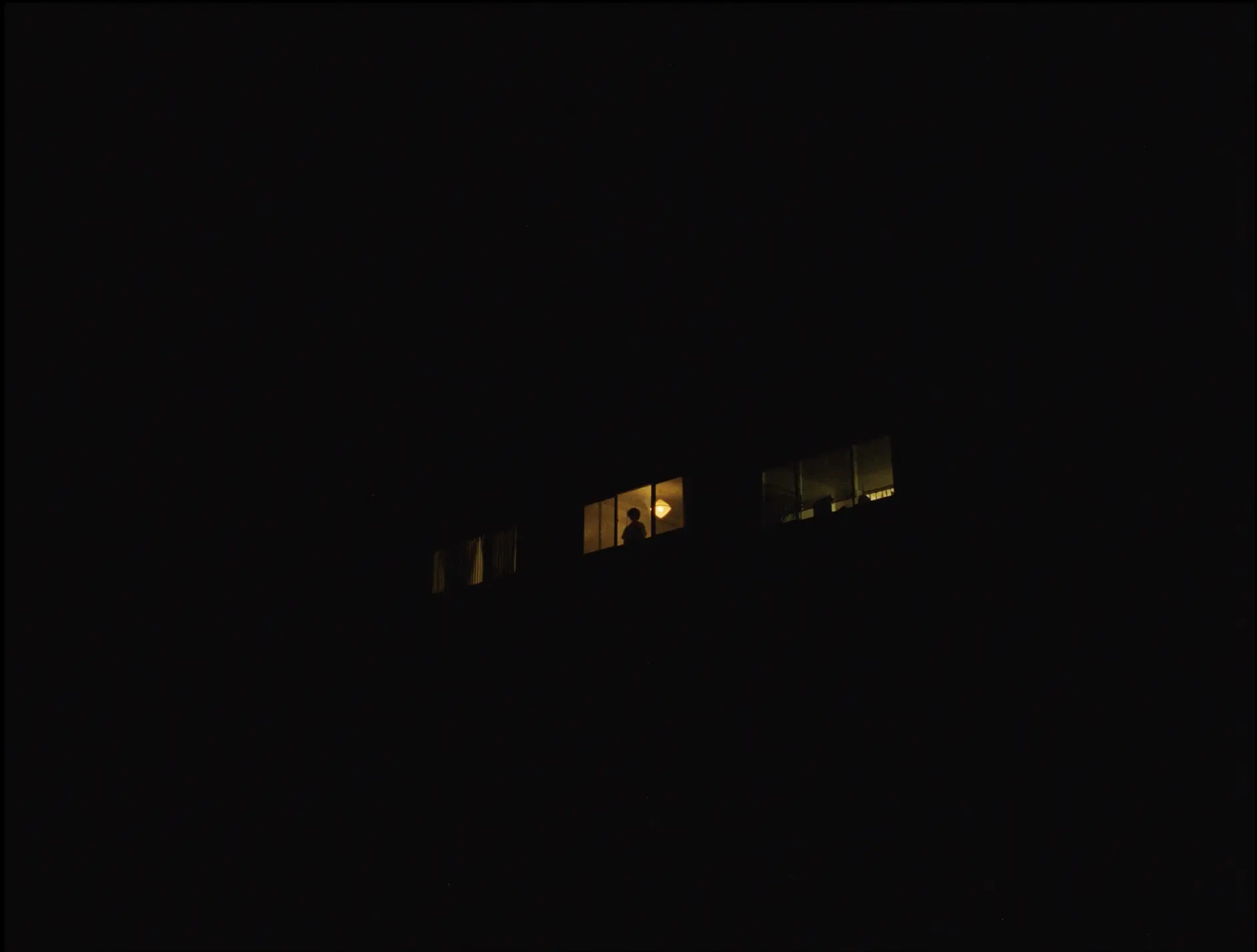 A distant window glowing with warm light in the darkness, showing a silhouette of a person standing inside.