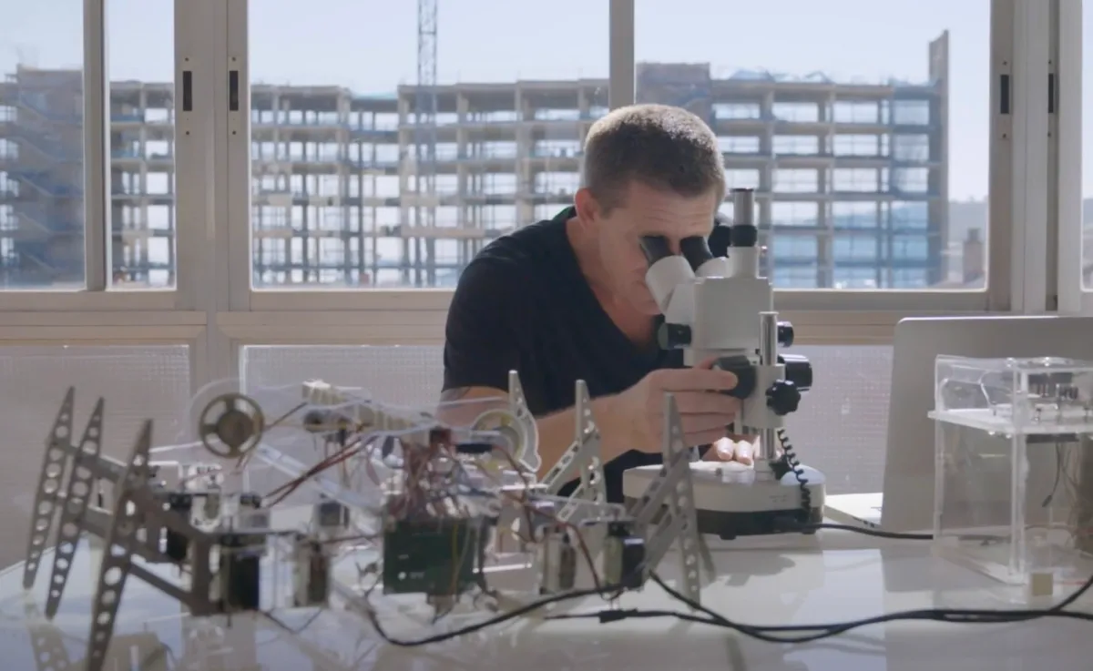 An individual in a laboratory peering into a microscope, with a machine prototype laid on the desk. Windows in the background offer a view of a building under construction.