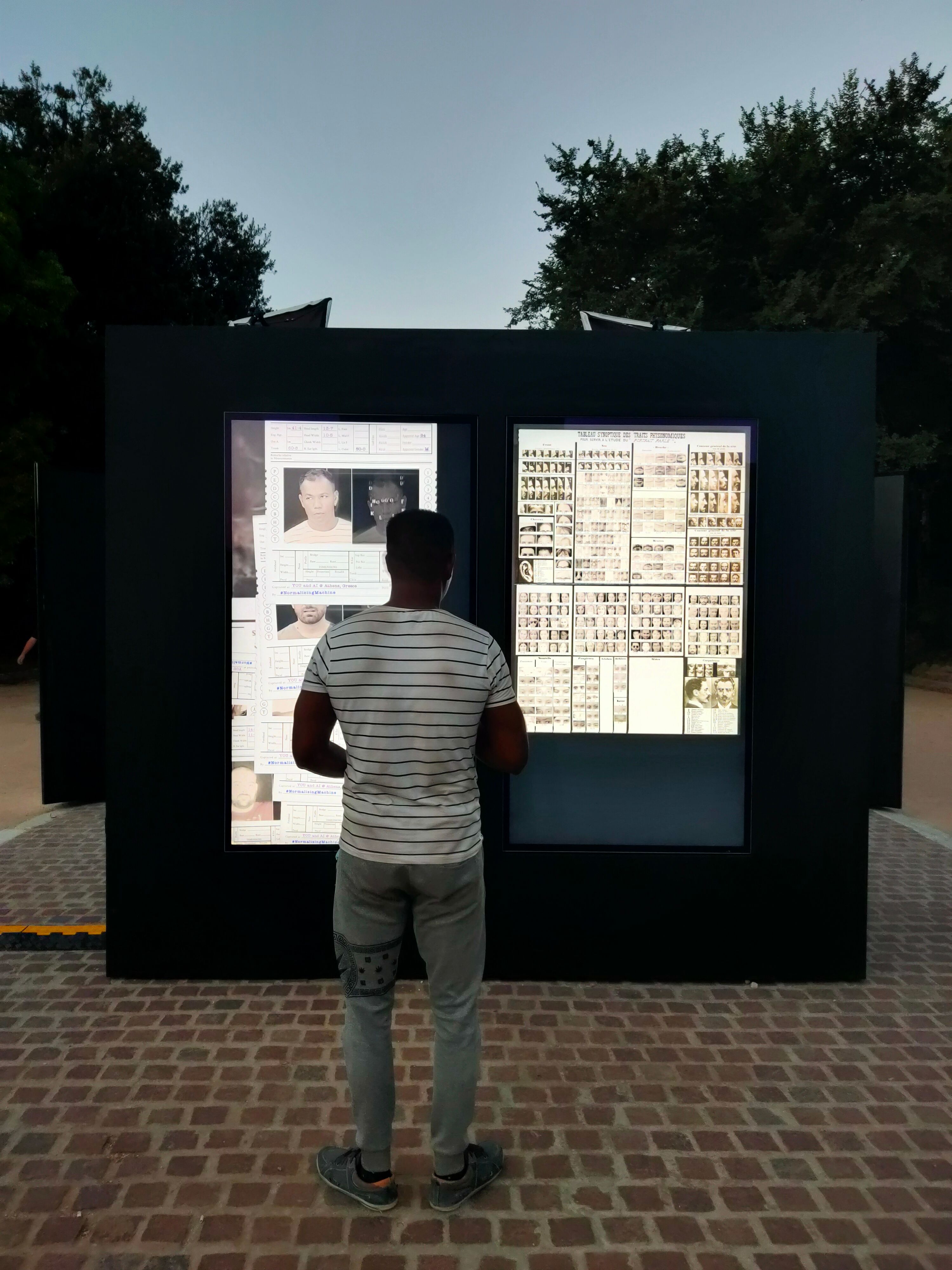 A person standing in front of a digital display featuring historical photographs and documents in an outdoor setting at dusk.
