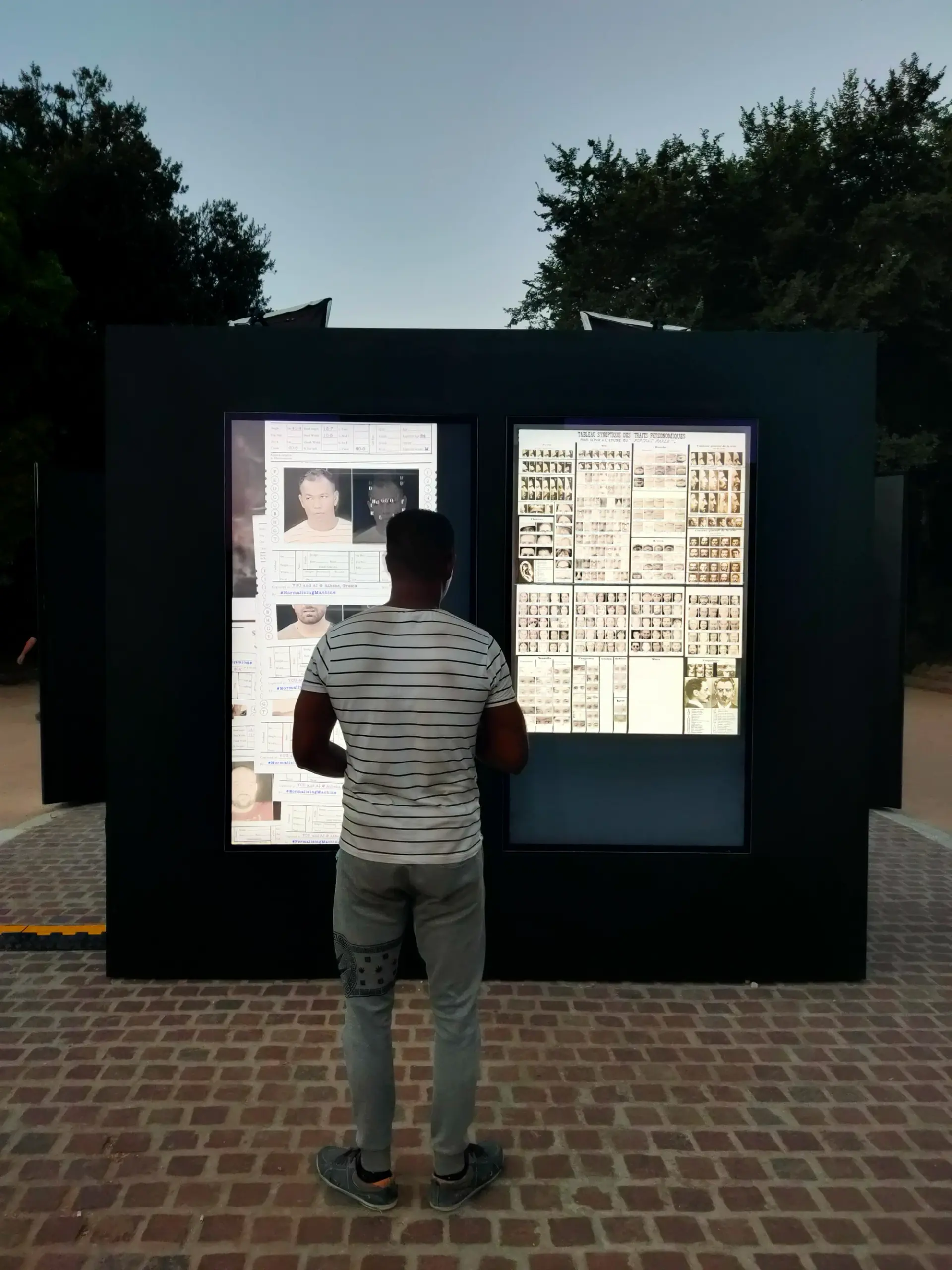 A person standing in front of a digital display featuring historical photographs and documents in an outdoor setting at dusk.