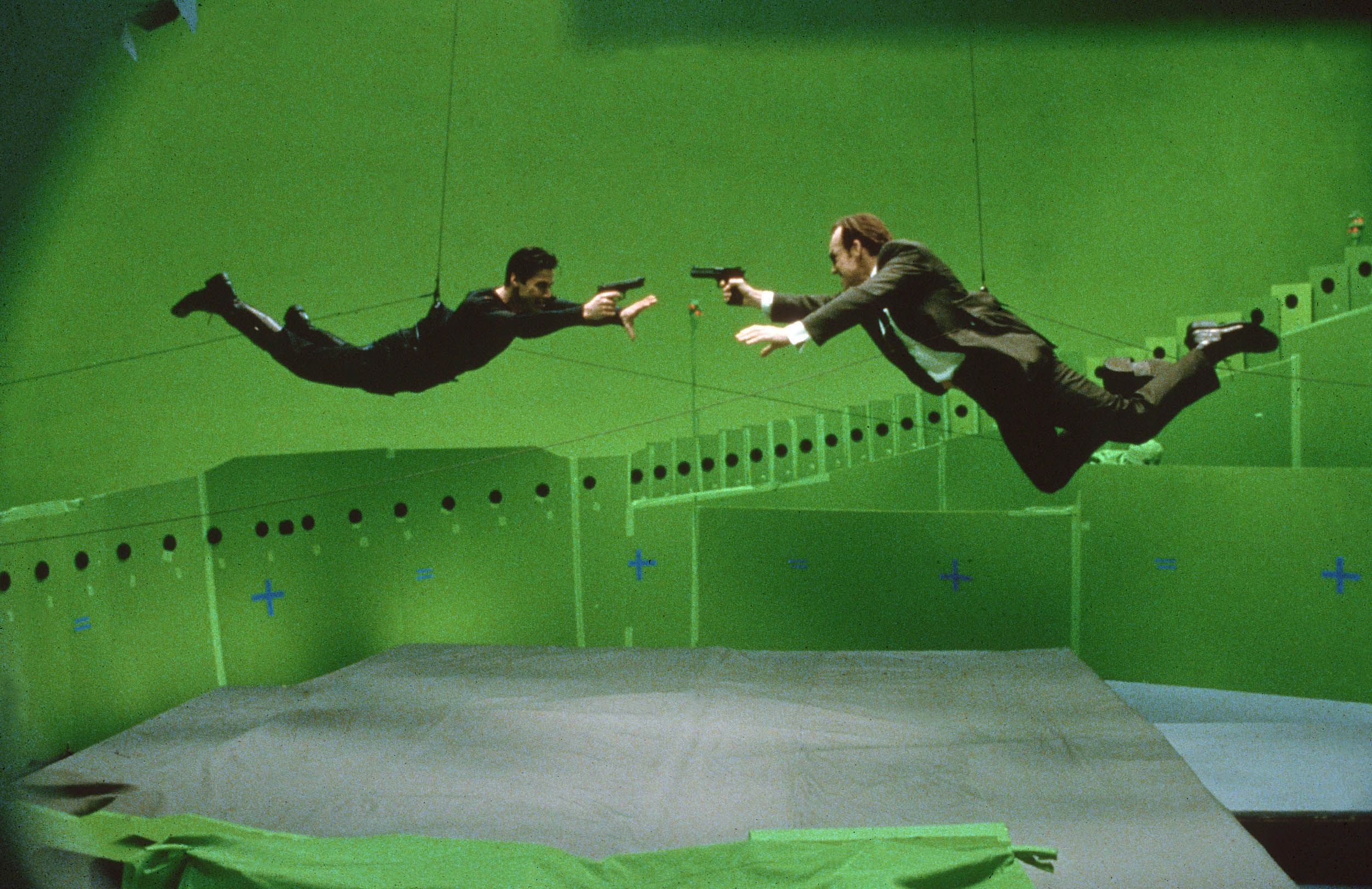 Actors in mid-air fight positions on wires, set against a green screen backdrop.