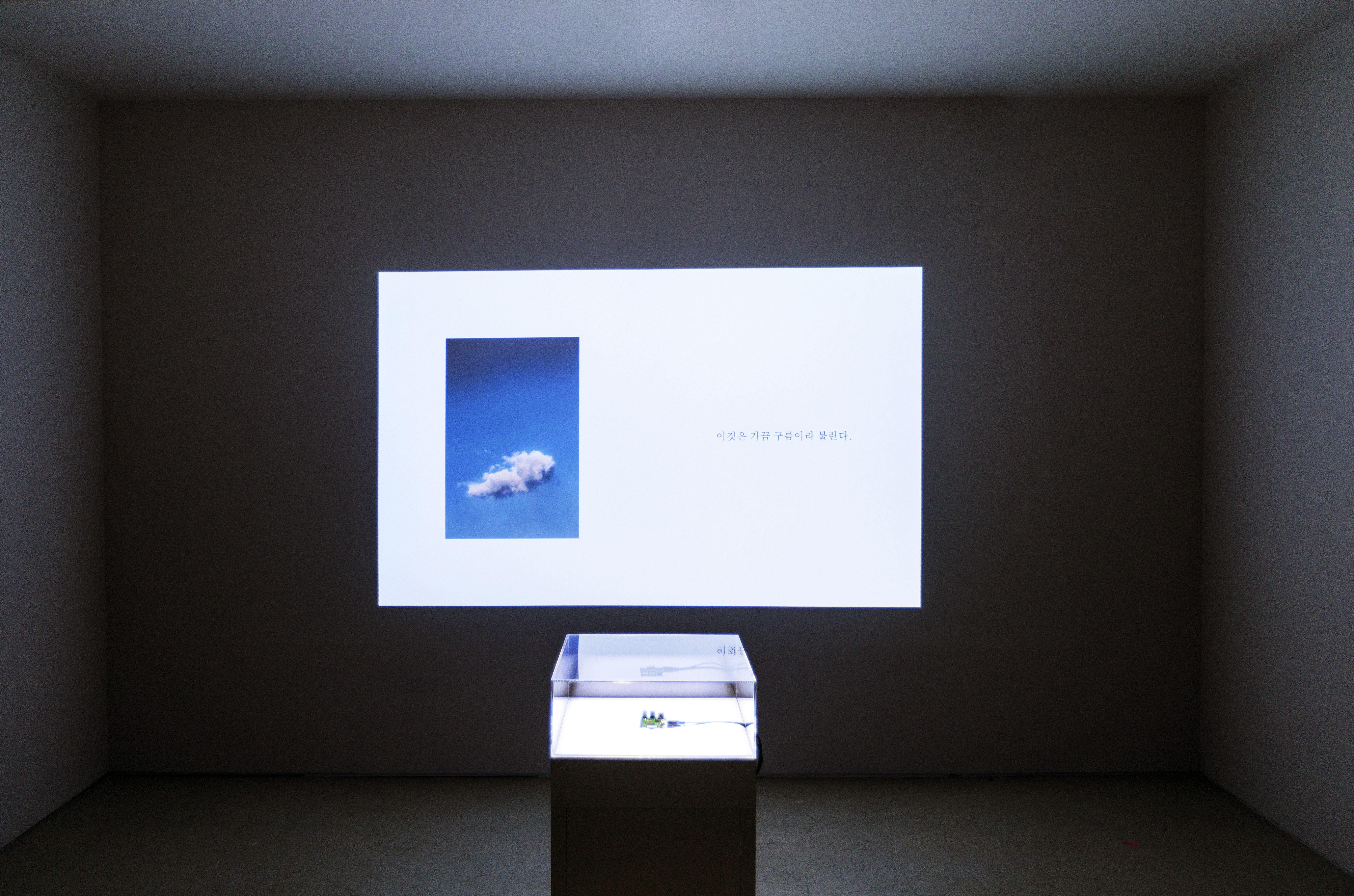 A transparent cube placed on a black pedestal in a dimly lit room, with an image projection on the wall behind it.