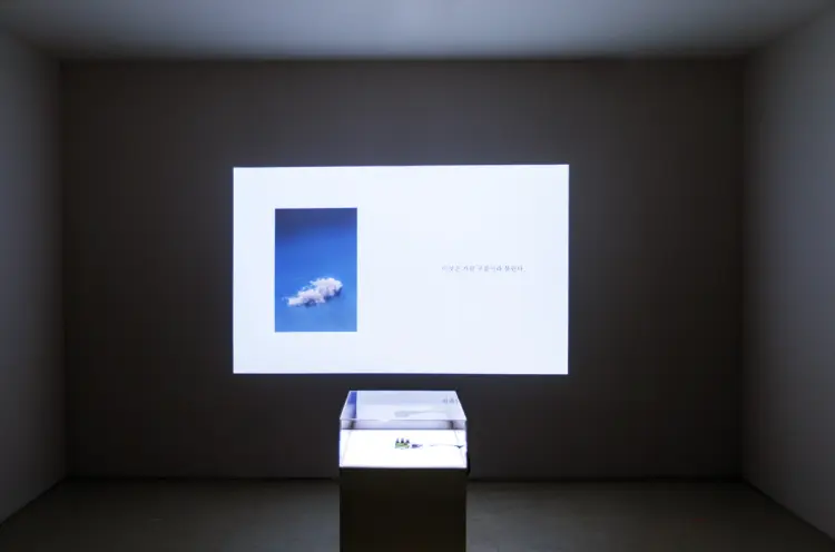 A transparent cube placed on a black pedestal in a dimly lit room, with an image projection on the wall behind it.