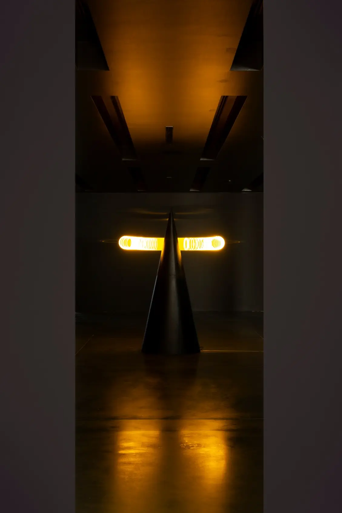 A symmetrical photograph captures a cone in a dark room radiating golden lights from its apex, which reflects captivatingly off its sides.