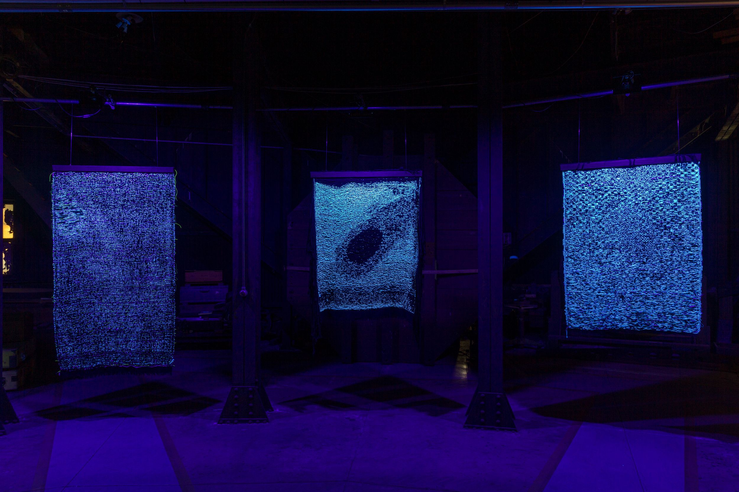 Three tapestry artworks displayed in a room bathed in soft blue light.