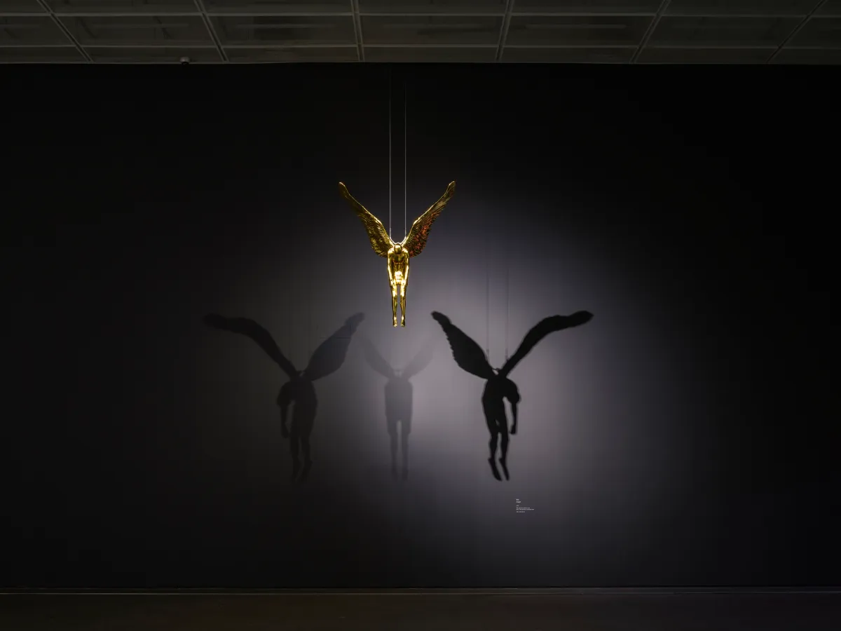 A golden angel statue suspended from the ceiling. The spotlight illuminates it, casting dramatic shadows on the black wall behind.
