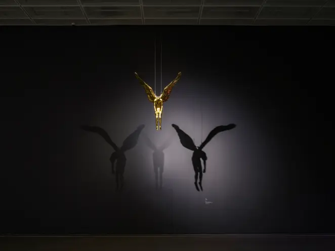 A golden angel statue suspended from the ceiling. The spotlight illuminates it, casting dramatic shadows on the black wall...
