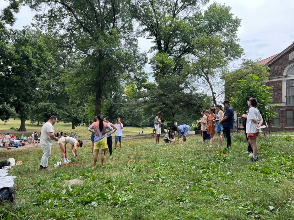 Group of people outdoors, some working with yarn on the ground, others observing, in a park setting.