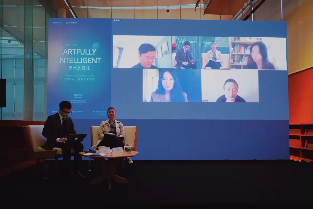 Two individuals are seated in front of a large screen projecting a virtual meeting. The screen content is featured within the context of the "Artfully Intelligent" exhibition at Yuz Museum.