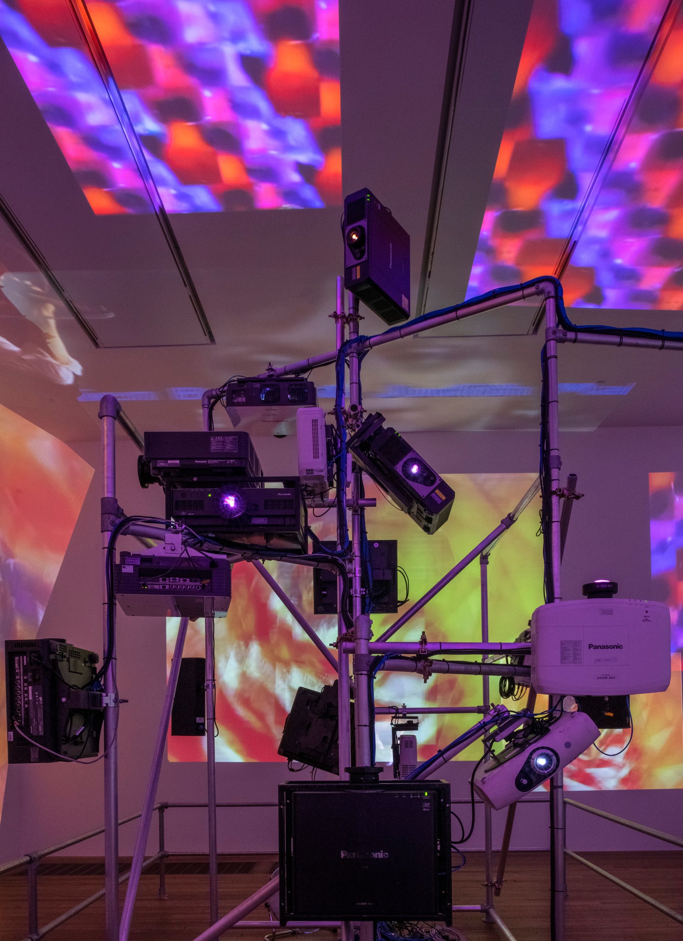 Projection equipment and pipes in an indoor setting, casting vibrant yellow, blue, and red light projections onto the ceil...