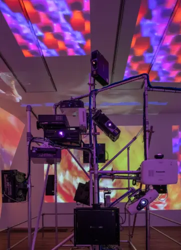 Projection equipment and pipes in an indoor setting, casting vibrant yellow, blue, and red light projections onto the ceil...