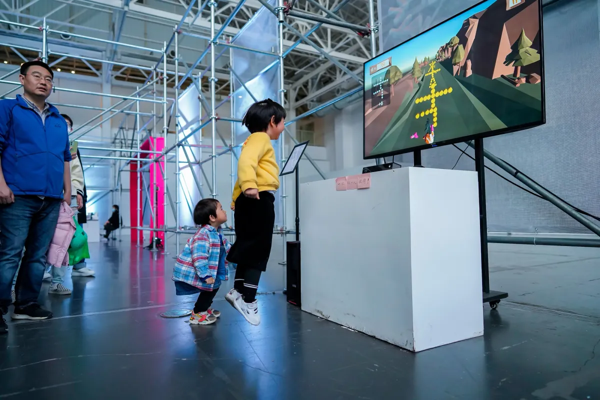 A gallery hall displaying a game on a monitor, with children energetically jumping in front of the screen. Some adults are standing behind them, watching the activity.