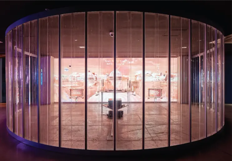 A round room with glass walls features a large screen positioned in front of a desk.