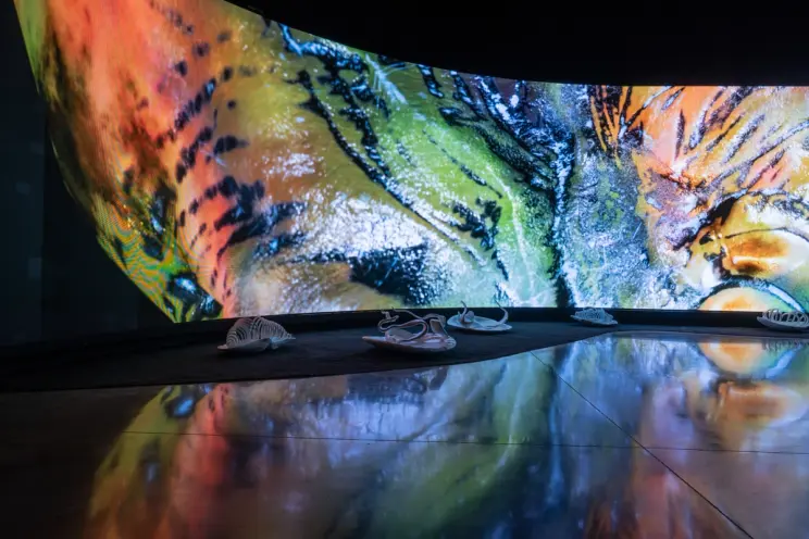 Curved digital display with colorful patterns and sneakers on reflective floor.