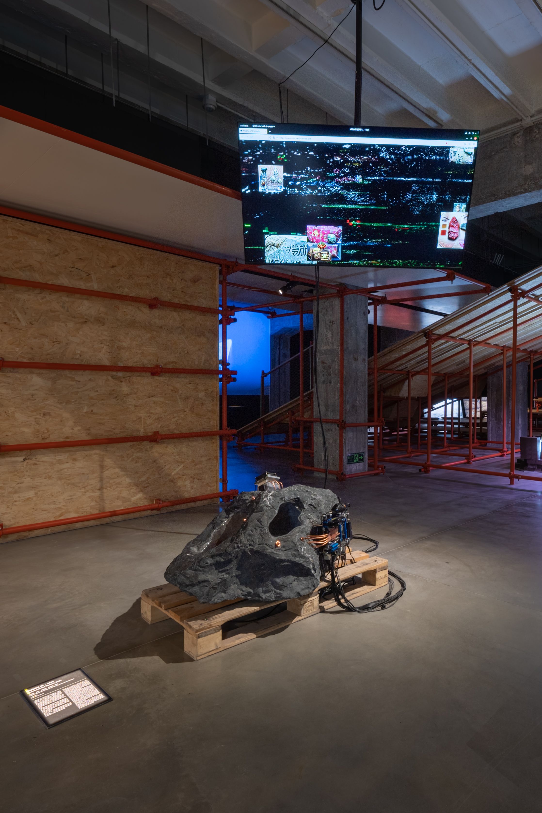 Rock-like sculpture with electronics on a pallet, screen in the background.