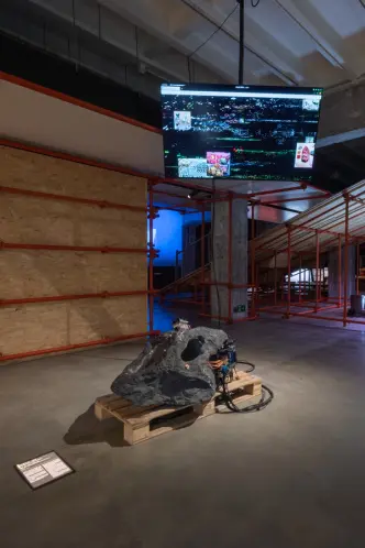 Rock-like sculpture with electronics on a pallet, screen in the background.