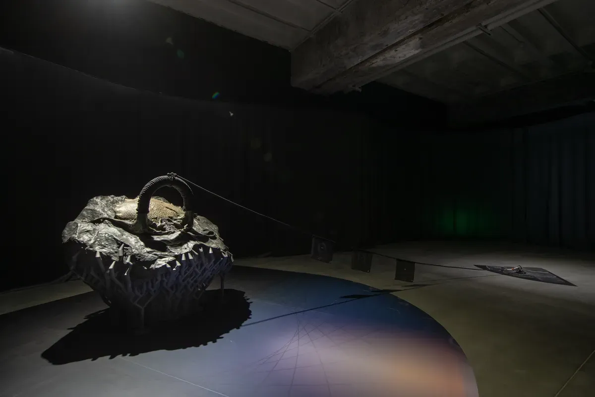 A rugged, textured sculpture resembling a large rock sits in a dimly lit gallery space. A rope is firmly attached to it, suggesting interaction or potential movement.