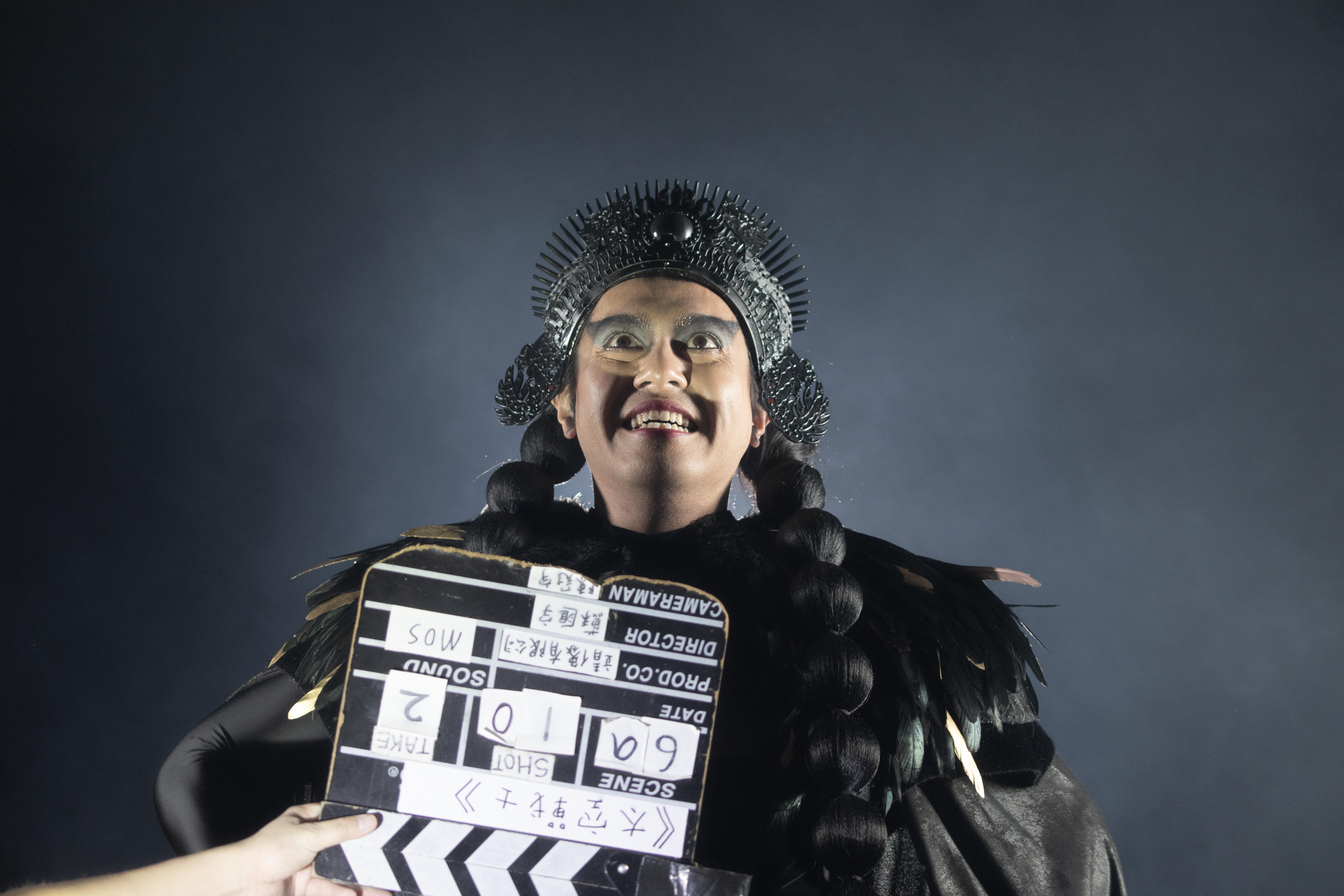An individual donned in a black feathered coat and an iron crown stands in front of a director's cut board, their face upt...