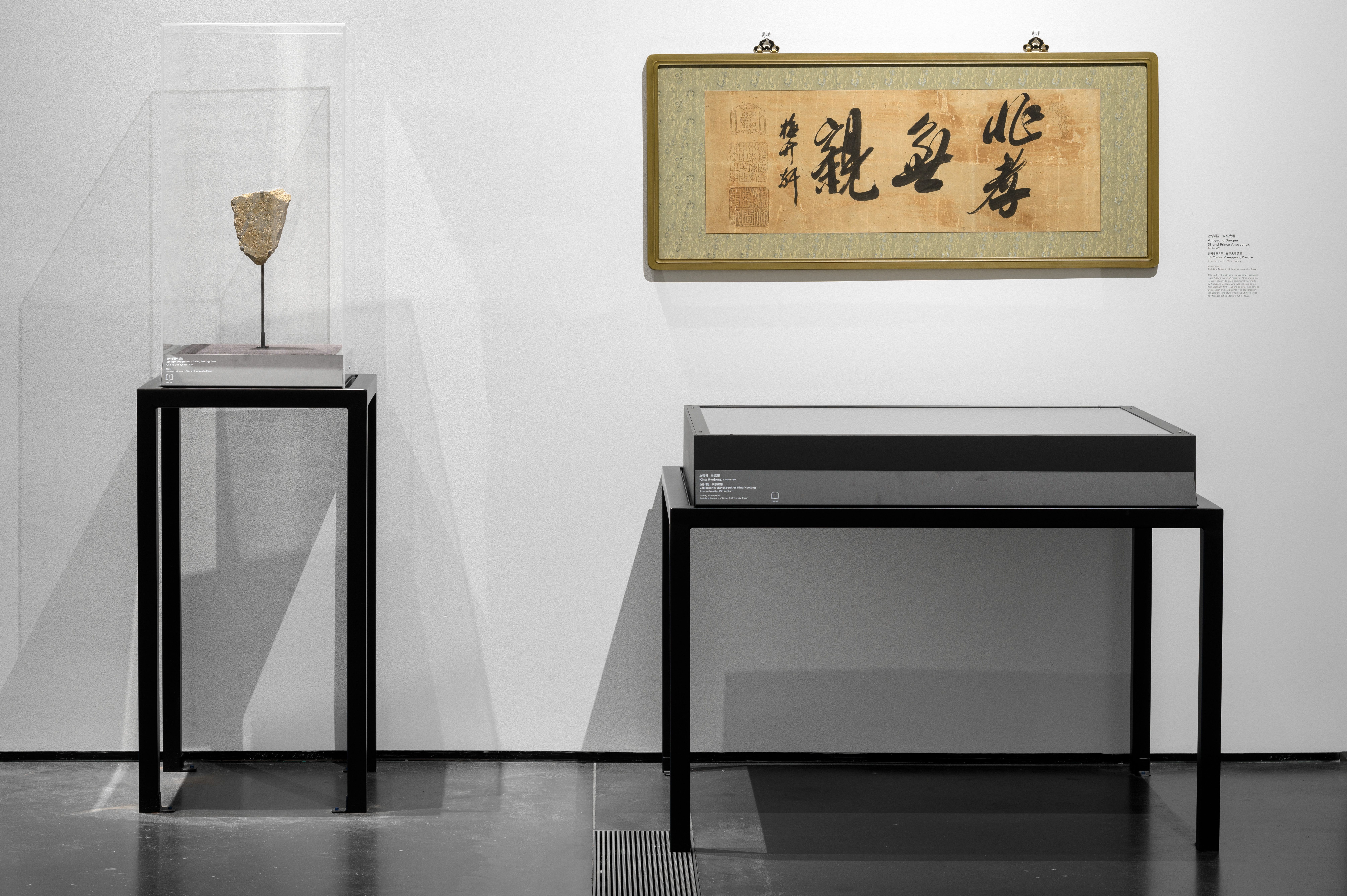 Several art pieces displayed on metallic stands, contrasting against a white wall and black floor. One artwork is hung pro...