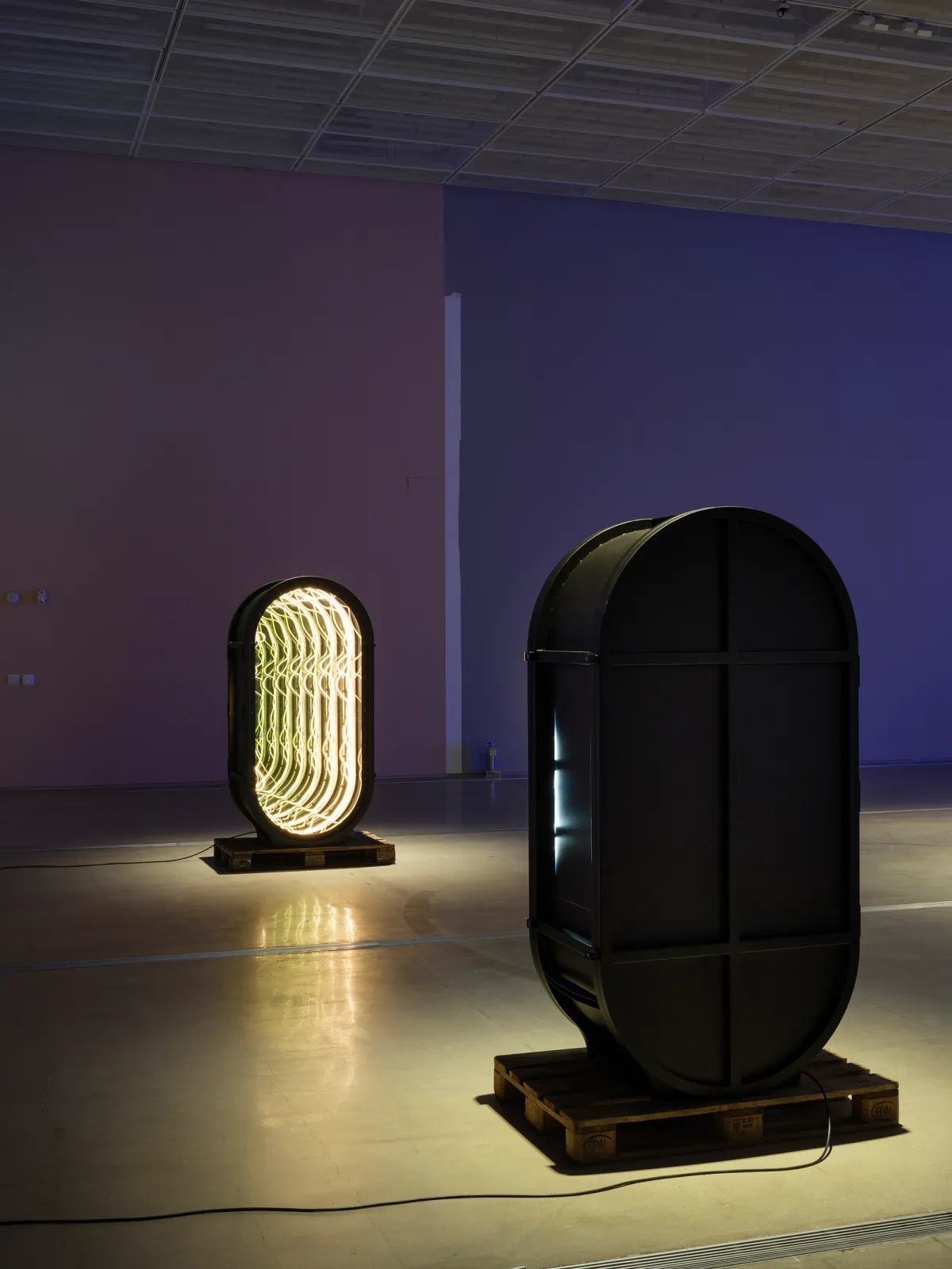 Two device-like sculptures resembling portals, stand independently in an art installation room with purple walls.