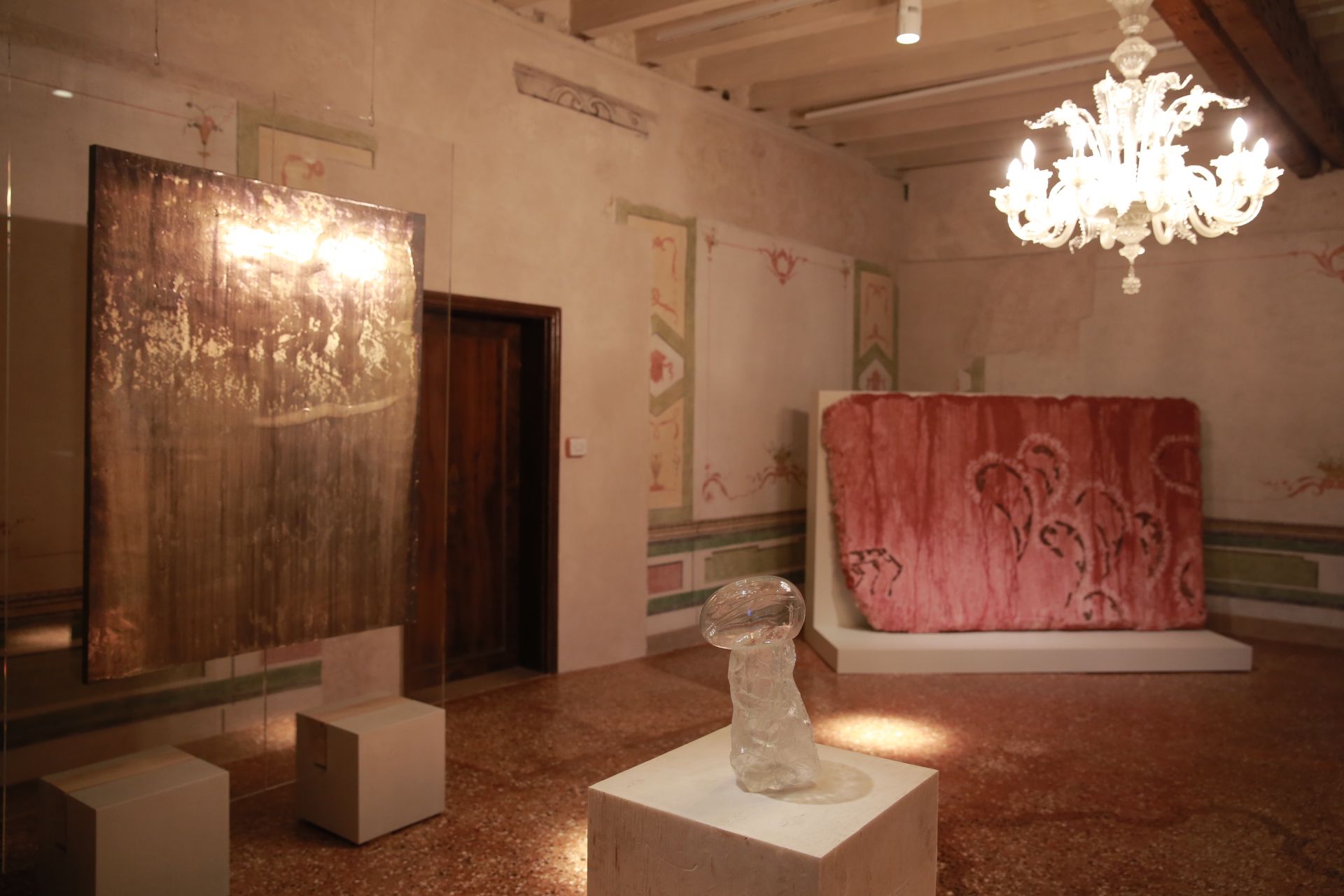 Warmly lit gallery with a metallic piece, glass sculpture, textile art, and ornate glass chandelier.