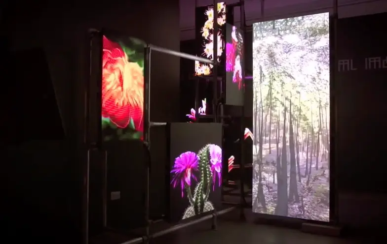 An art installation showcasing multiple screens attached to black pipes in a dark room with black walls. The screens displ...