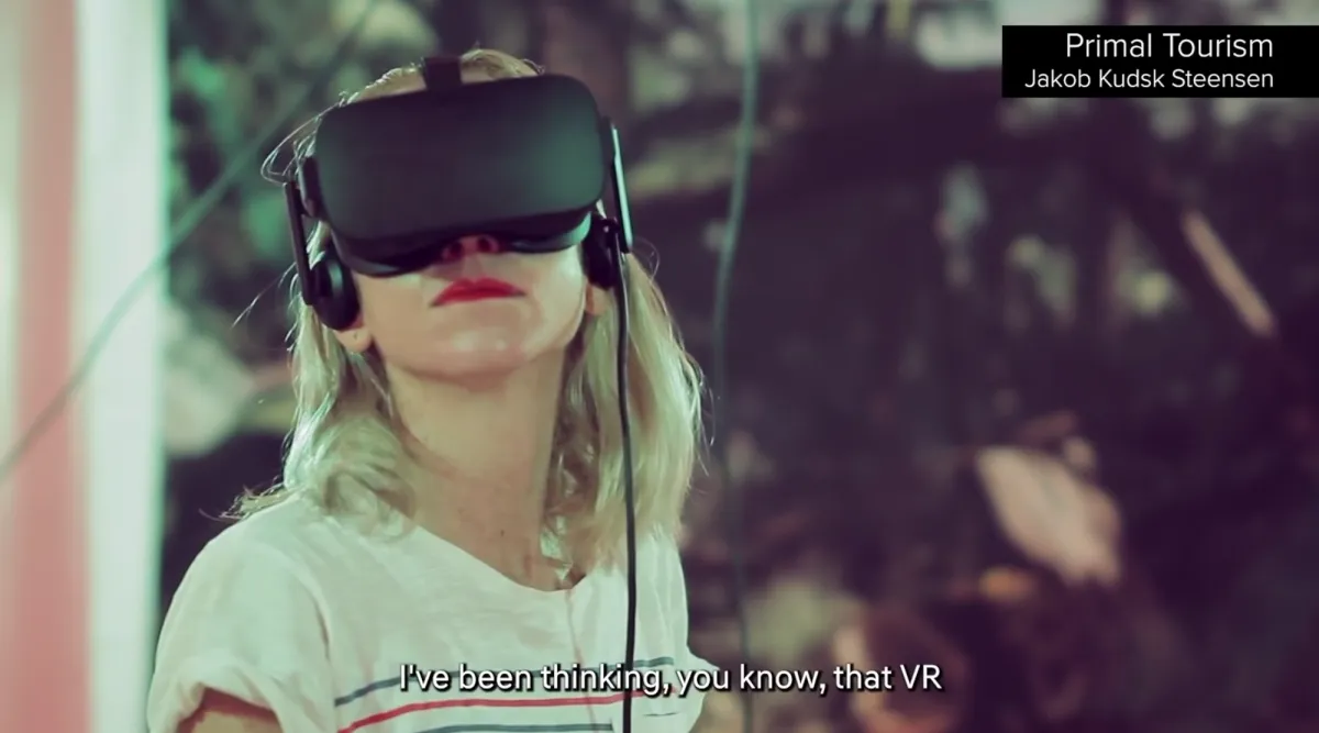 A blond individual wearing a black virtual reality headset and headphones. Their red lipstick stands out against their pale skin. The background features blurred nature imagery. The text in the upper right notes "Primal Tourism, Jakob Kudsk Steensen", and the text at the bottom starts with "I've been thinking, you know, that VR".