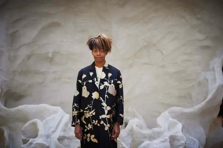 A person in floral patterned formal clothes stands before an enormous white statue inside a spacious gallery.